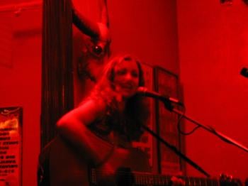 Woman playing guitar under red light, with a glowing-eye skull behind her head, at a bar on the Upper East Side.