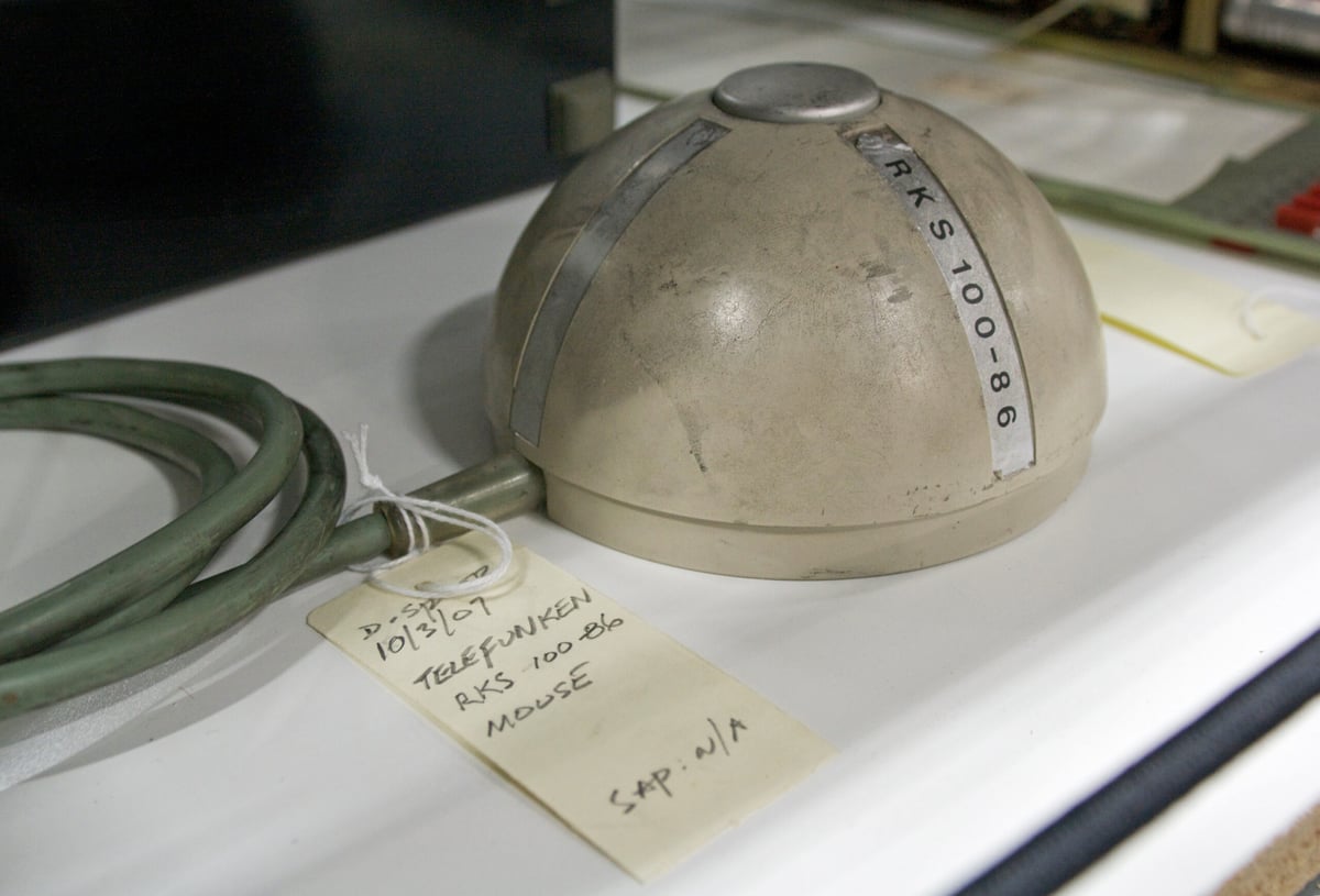 Photograph of an early rolling-ball mouse from the 1960s, shaped like half a cylinder and sitting on a desk with a label.