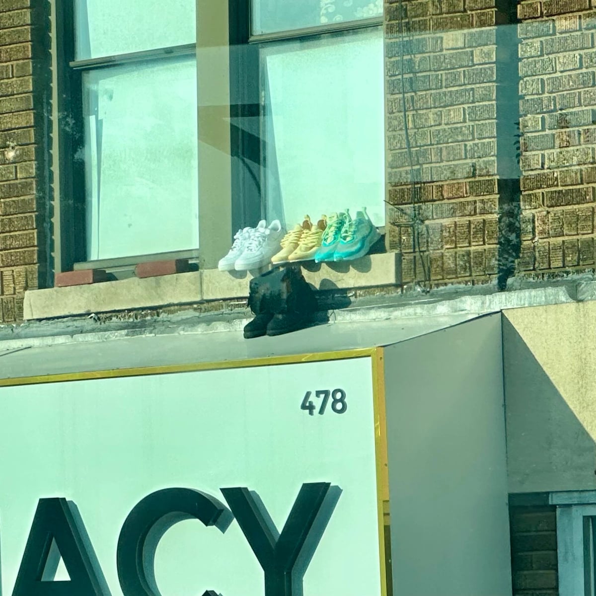 Boots and three pairs of sneakers on a window ledge. The sneakers are white, yellow, and turquoise. The boots are black and are placed right on the metal awning of the pharmacy below.