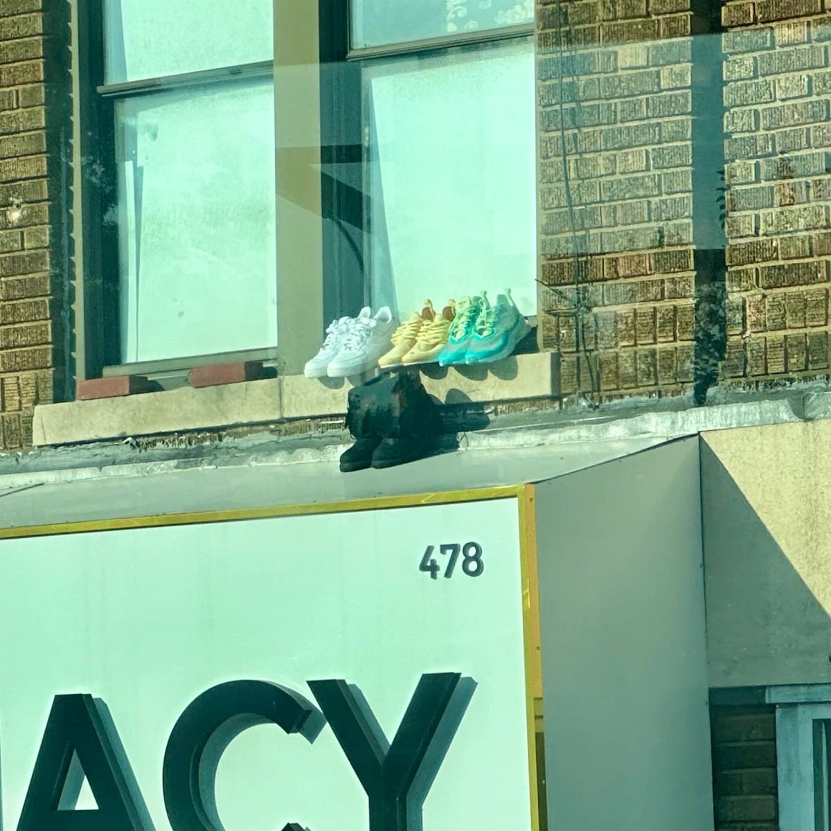 Boots and three pairs of sneakers on a window ledge. The sneakers are white, yellow, and turquoise. The boots are black and are placed right on the metal awning of the pharmacy below.