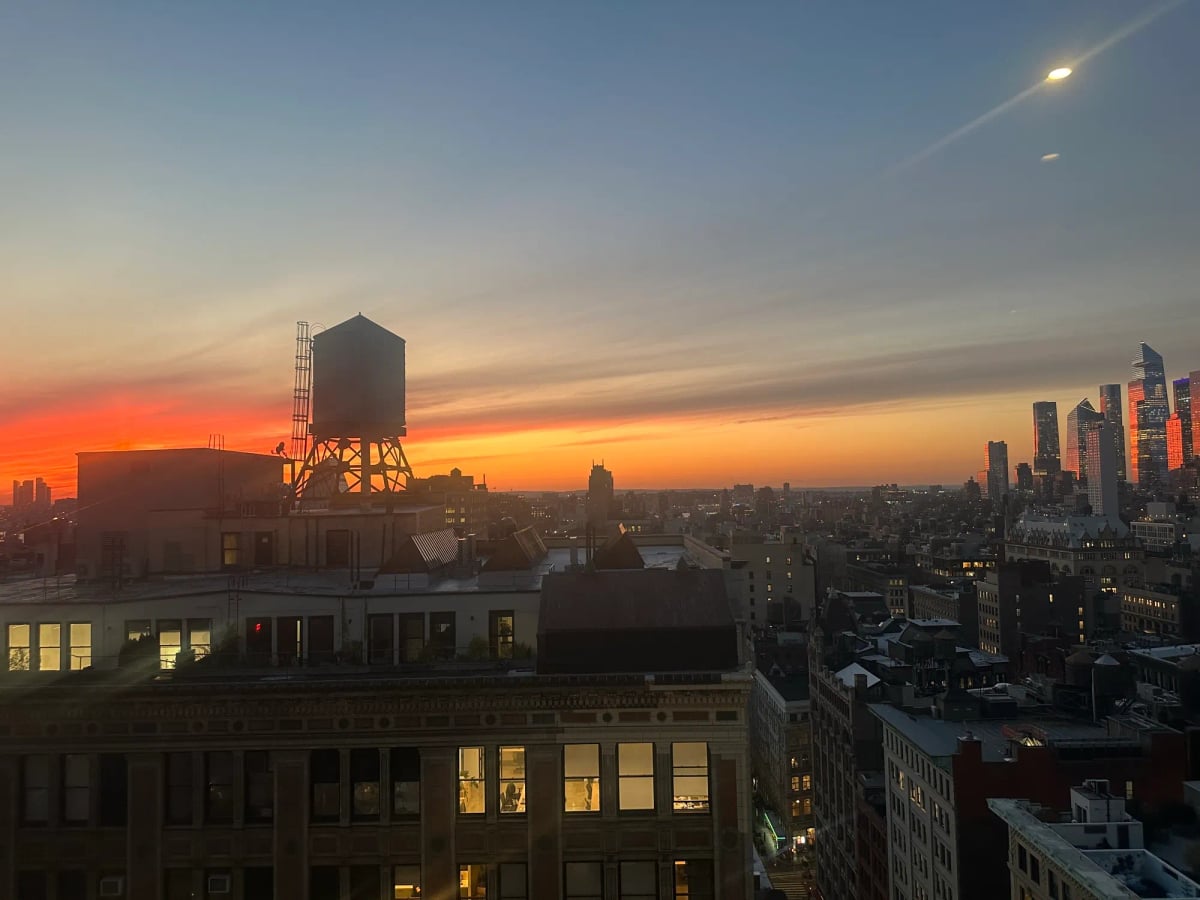 A view of the New York City skyline facing West, at sunset. The colors of the sunset, deep orange, and yellow, fill the sky behind the silhouette of a water tower atop a 20 story building, with some of the windows illuminated as people do their work into the evening. Avenues stretch towards the Hudson, and on the far right the cluster of buildings that make up Hudson yards glow suffused with the orange of the sunset. Even Hudson yards can be made beautiful, especially at a distance.