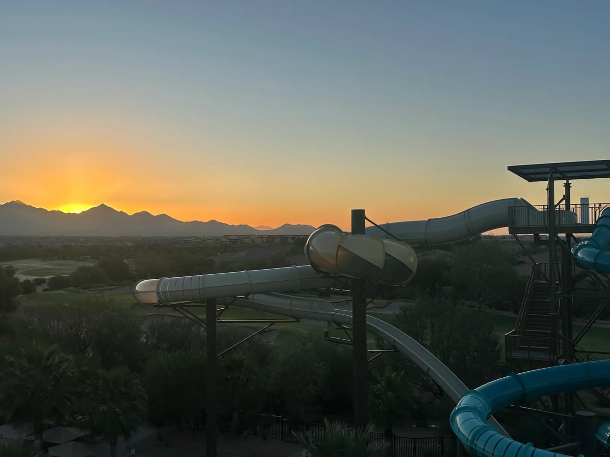 The sun rises over distant mountains—a beautiful desert scene. In the darker foreground a large looping water slide fills in half the frame on the right.