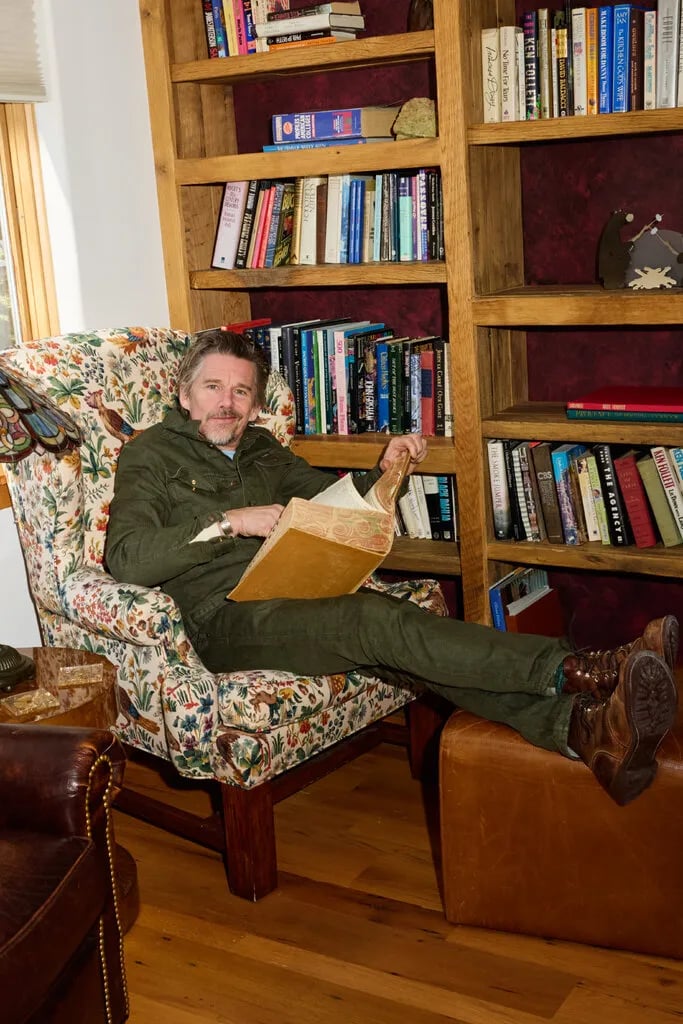 A smirking Ethan Hawke dressed all in military shades of green with boots on, in a hideous armchair upholstered with a bird and plants print, one of those things that is so relentlessly ugly it signifies sophistication and probably costs $9,000, in front of a bunch of books. He's reading a massive, marble-edged, leather-bound book that appears to be a dictionary. He pulls it off because he's just an earnest poetic sex angel and we all know it but literally anyone else would be dragged for eons through every tranche of hell.