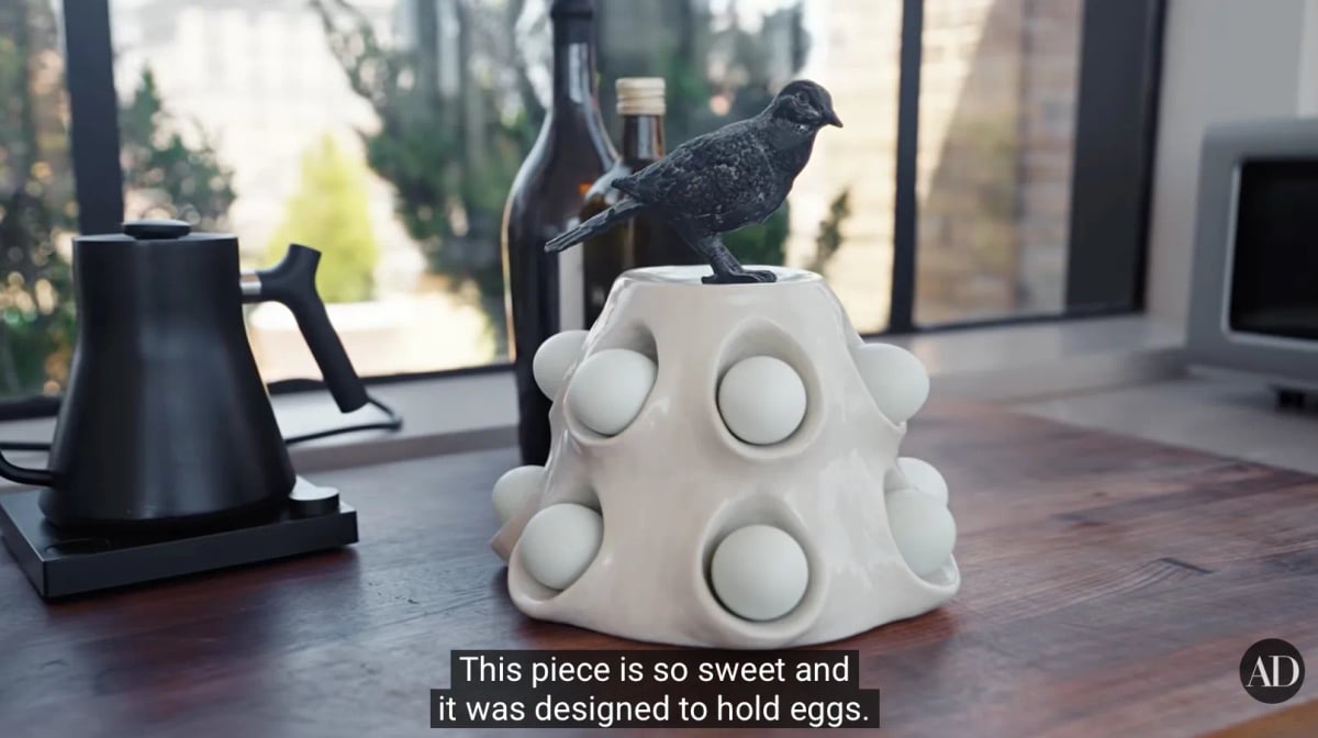 A knobby, vaguely pyramidal white ceramic object on a kitchen counter, perhaps a foot tall, with a black rough metal bird perched atop; the pyramidal ceramic has a series of pouchlike crevices with labial rims, nestling nine white eggs; those are the visible eggs. The caption reads "This piece is so sweet and it was designed to hold eggs." While the video does not directly address it, it is reasonable to assume that this is how new Julianne Moores are incubated.