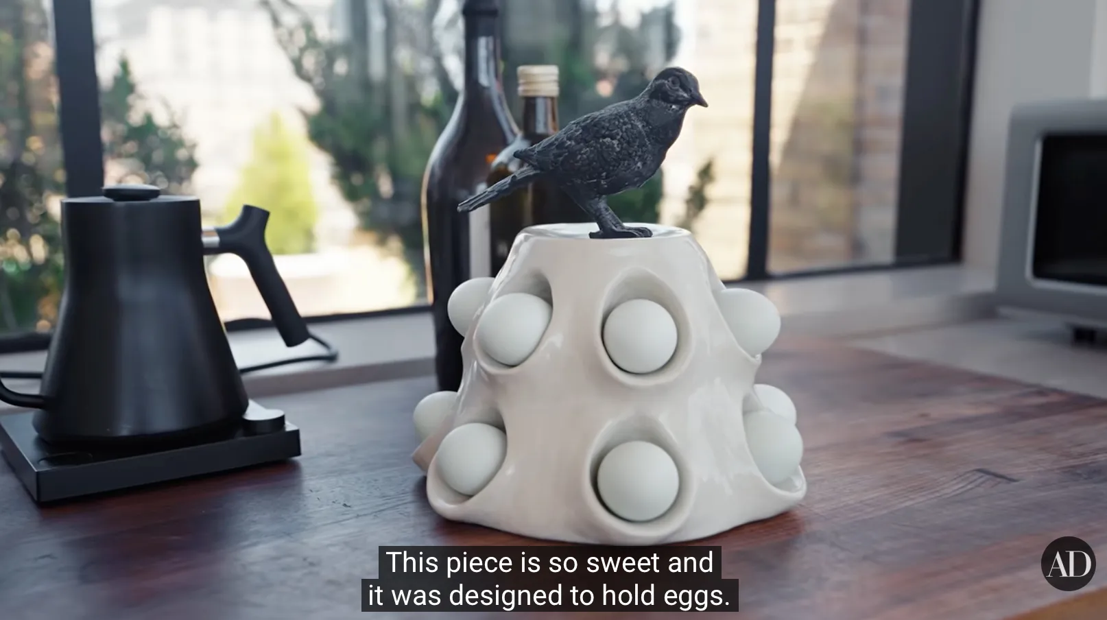 A knobby, vaguely pyramidal white ceramic object on a kitchen counter, perhaps a foot tall, with a black rough metal bird perched atop; the pyramidal ceramic has a series of pouchlike crevices with labial rims, nestling nine white eggs; those are the visible eggs. The caption reads "This piece is so sweet and it was designed to hold eggs." While the video does not directly address it, it is reasonable to assume that this is how new Julianne Moores are incubated.